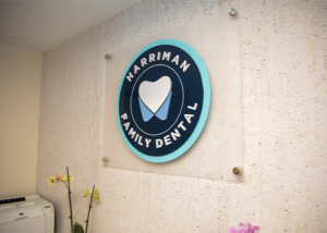 Wall-mounted circular sign for Harriman Family Dental featuring a white tooth icon flanked by blue abstract shapes on a teal background. The sign is secured with metallic fasteners above a printer and a vase of yellow and pink orchids in a reception area.