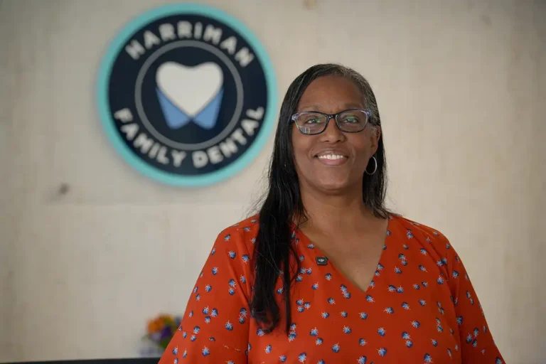 Individual in glasses and red patterned top standing indoors in front of circular Harriman Family Dental logo with heart design.